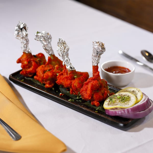 Four chicken lollipops with foil-wrapped bones, served with sauce, lemon, and red onion on a black plate.