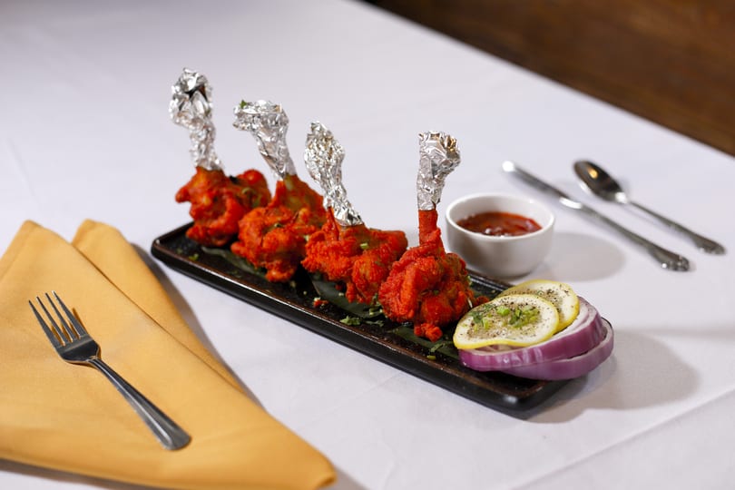 Four chicken lollipops with foil-wrapped bones, served with sauce, lemon, and red onion on a black plate.
