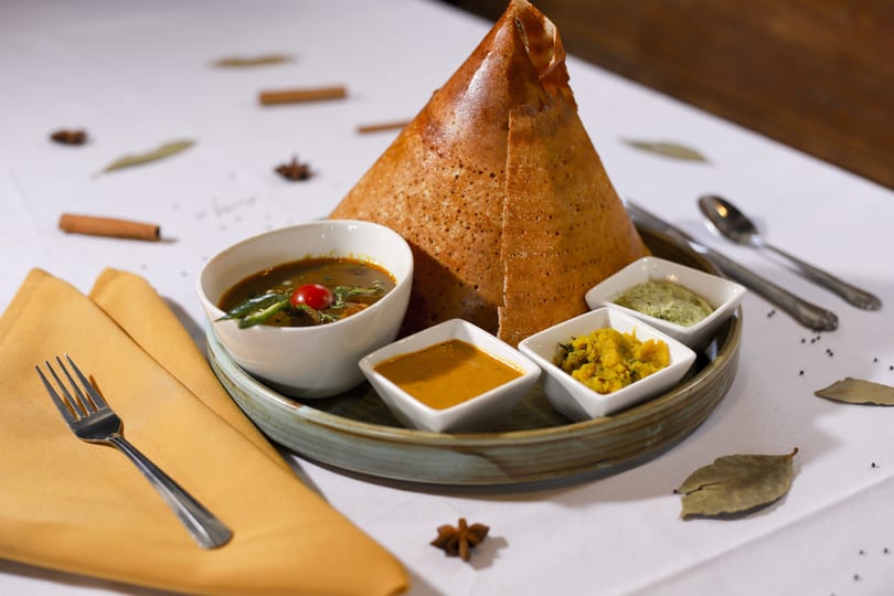 Crispy golden dosa, shaped like a cone, served with three small bowls of chutneys and a bowl of sambar.