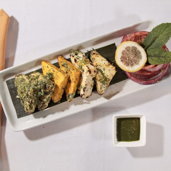 A white rectangular plate with grilled paneer, green chutney, lemon, and red onion on a white tablecloth.