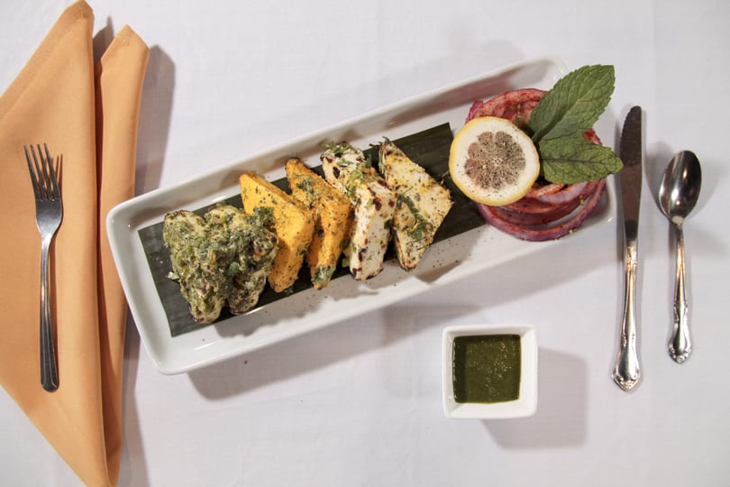 A white rectangular plate with grilled paneer, green chutney, lemon, and red onion on a white tablecloth.