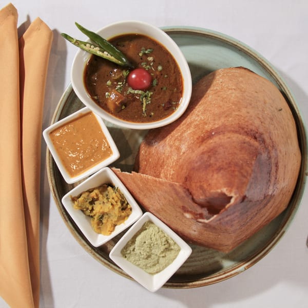 A dosa with three dipping sauces and a bowl of curry, garnished with chili and tomato.