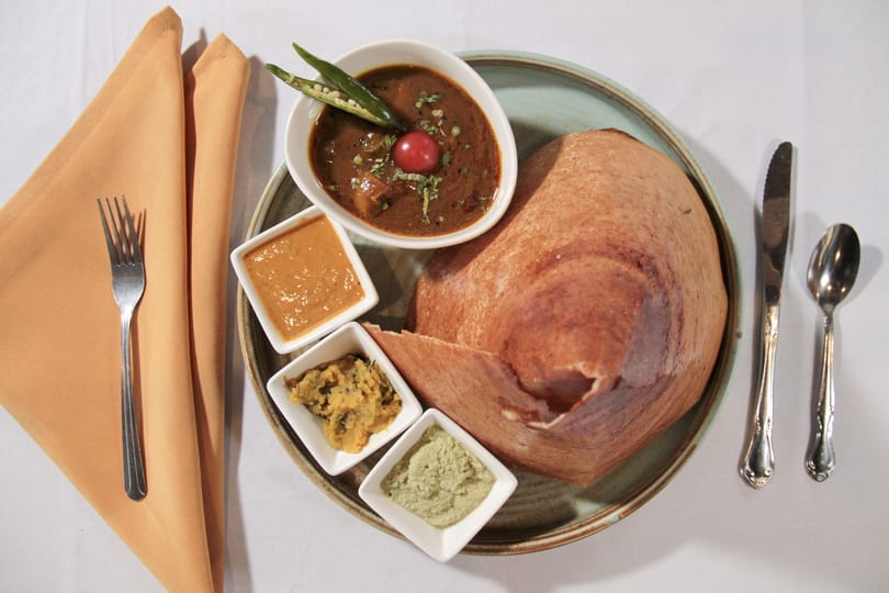 A dosa with three dipping sauces and a bowl of curry, garnished with chili and tomato.