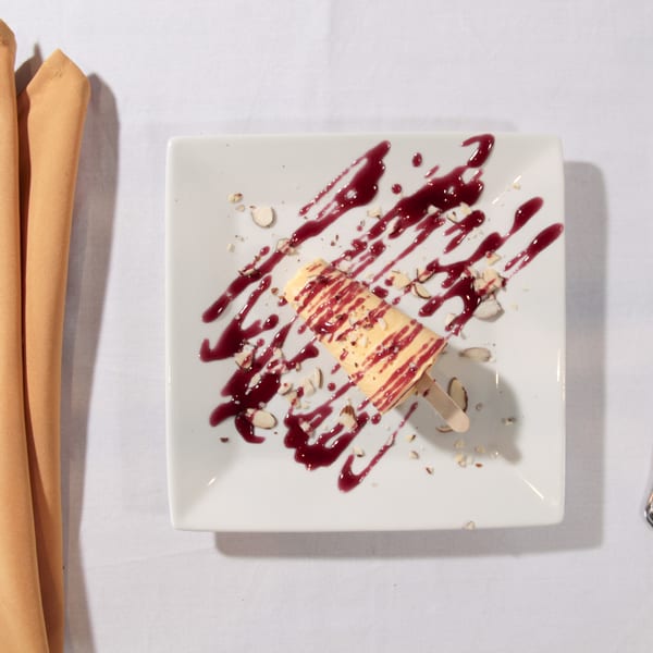 A dessert popsicle with berry sauce and nuts on a white plate, with cutlery and napkin.