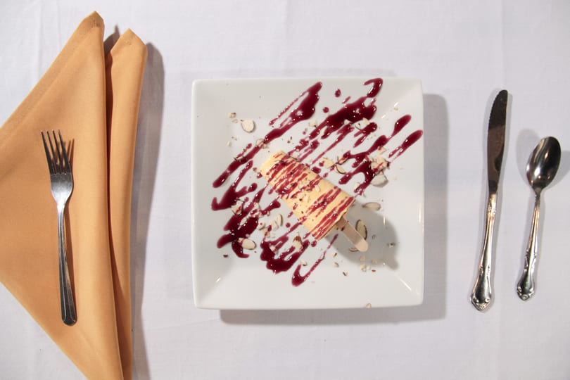 A dessert popsicle with berry sauce and nuts on a white plate, with cutlery and napkin.