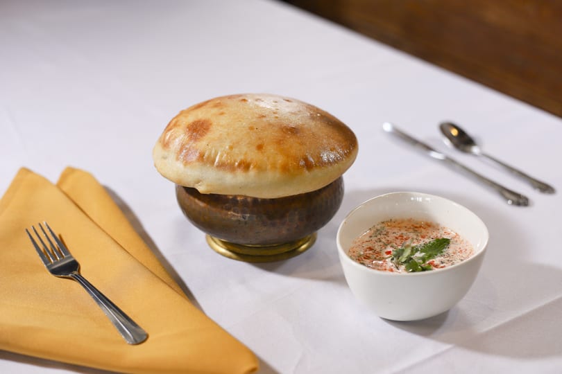 A golden-brown bread dome covers a copper pot, with a small bowl of raita and cutlery nearby.