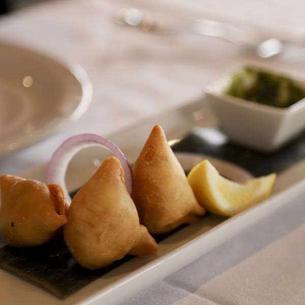 Three golden-brown samosas with red onion, lemon, and green chutney on a white rectangular plate.