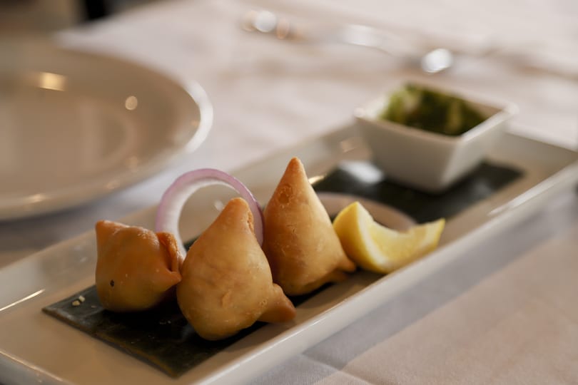 Three golden-brown samosas with red onion, lemon, and green chutney on a white rectangular plate.