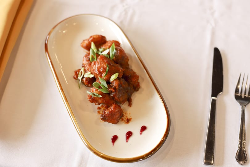 A plate of saucy chicken bites garnished with green onions and three drops of red sauce.